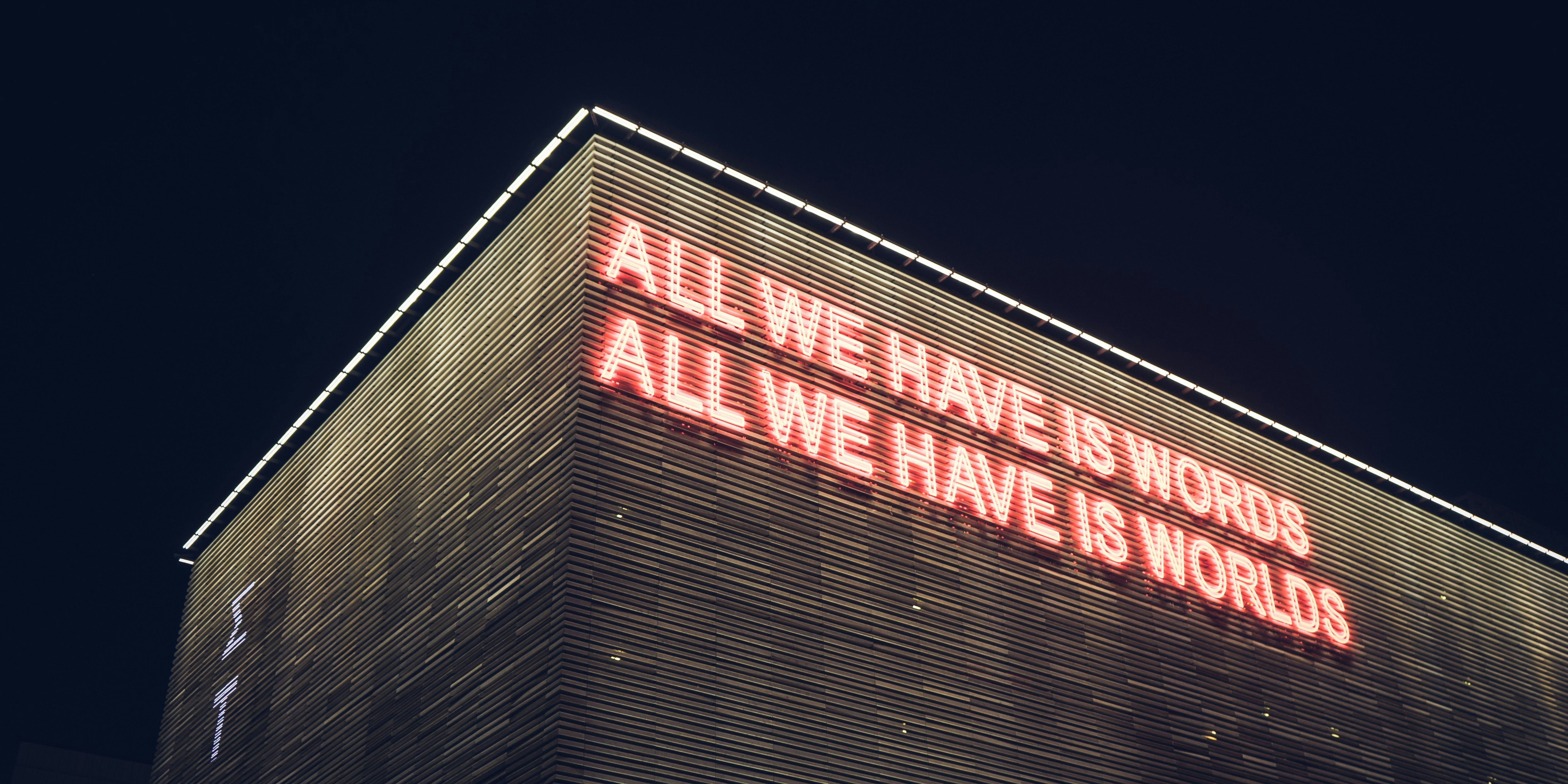 The illuminated facade of the Onassis Stegi building at night with the phrase "ALL WE HAVE IS WORDS" displayed.